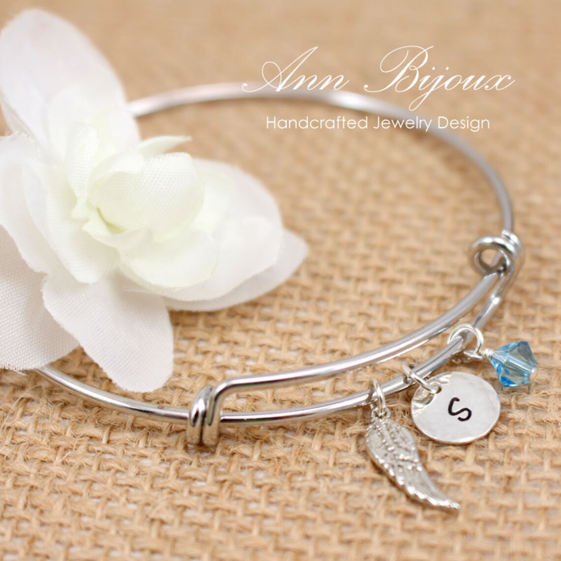 Ann Bijoux handcrafted jewelry design features a silver bracelet with an engraved initial 'S', angel wing charm, and a Swarovski birthstone accent.