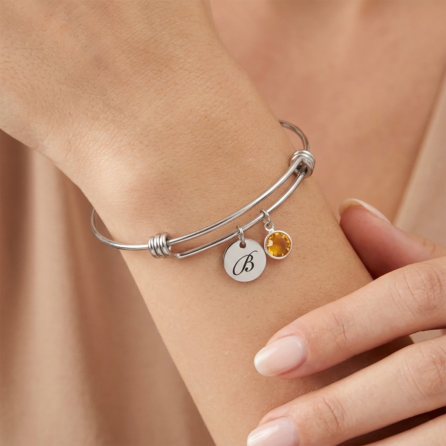Silver bracelet with a yellow gemstone and round charm on a wrist against a beige background