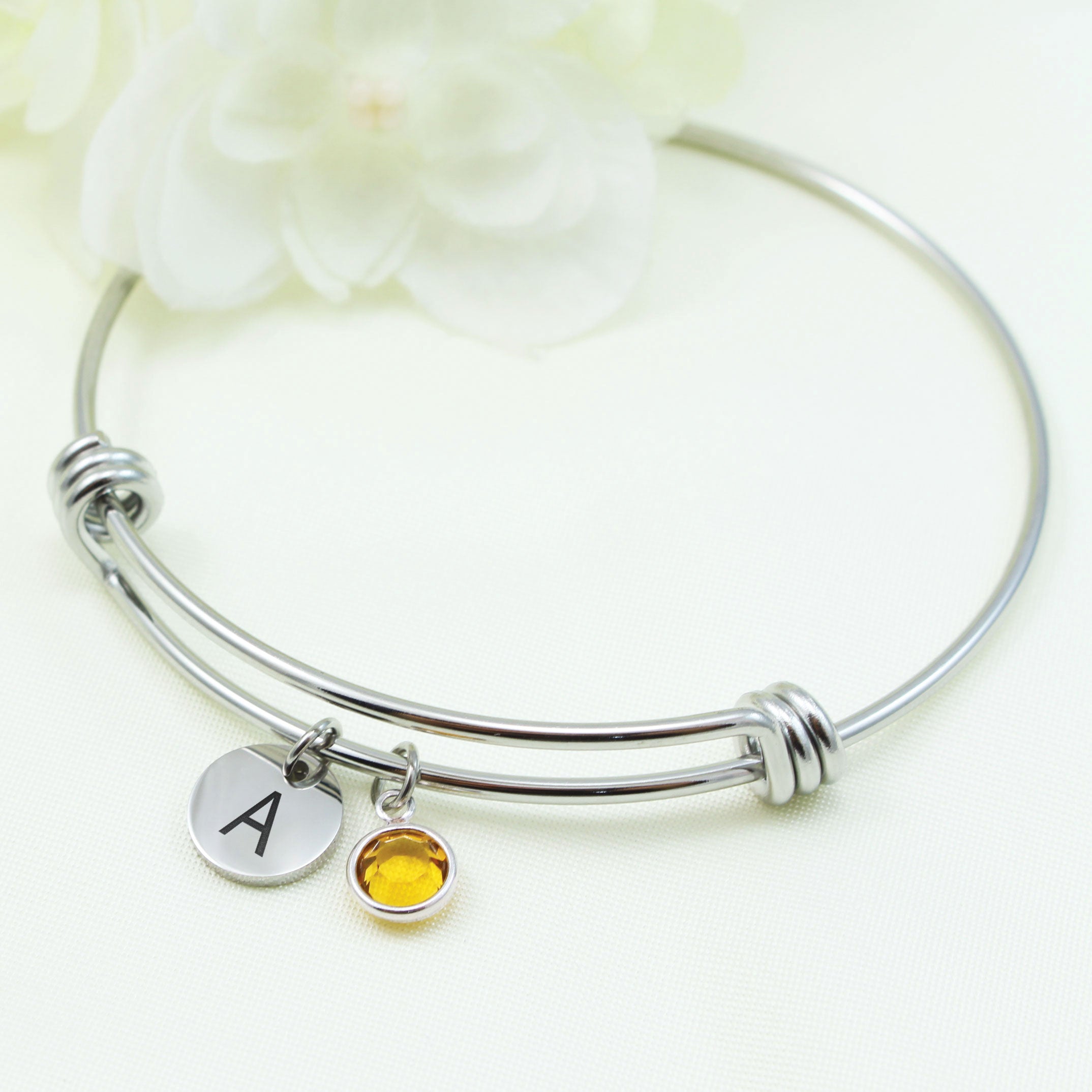 Close-up of a silver adjustable wire bangle on a wrist, featuring two hanging charms: a small silver disc engraved with the letter B in script and a circular amber-colored stone in a silver setting.