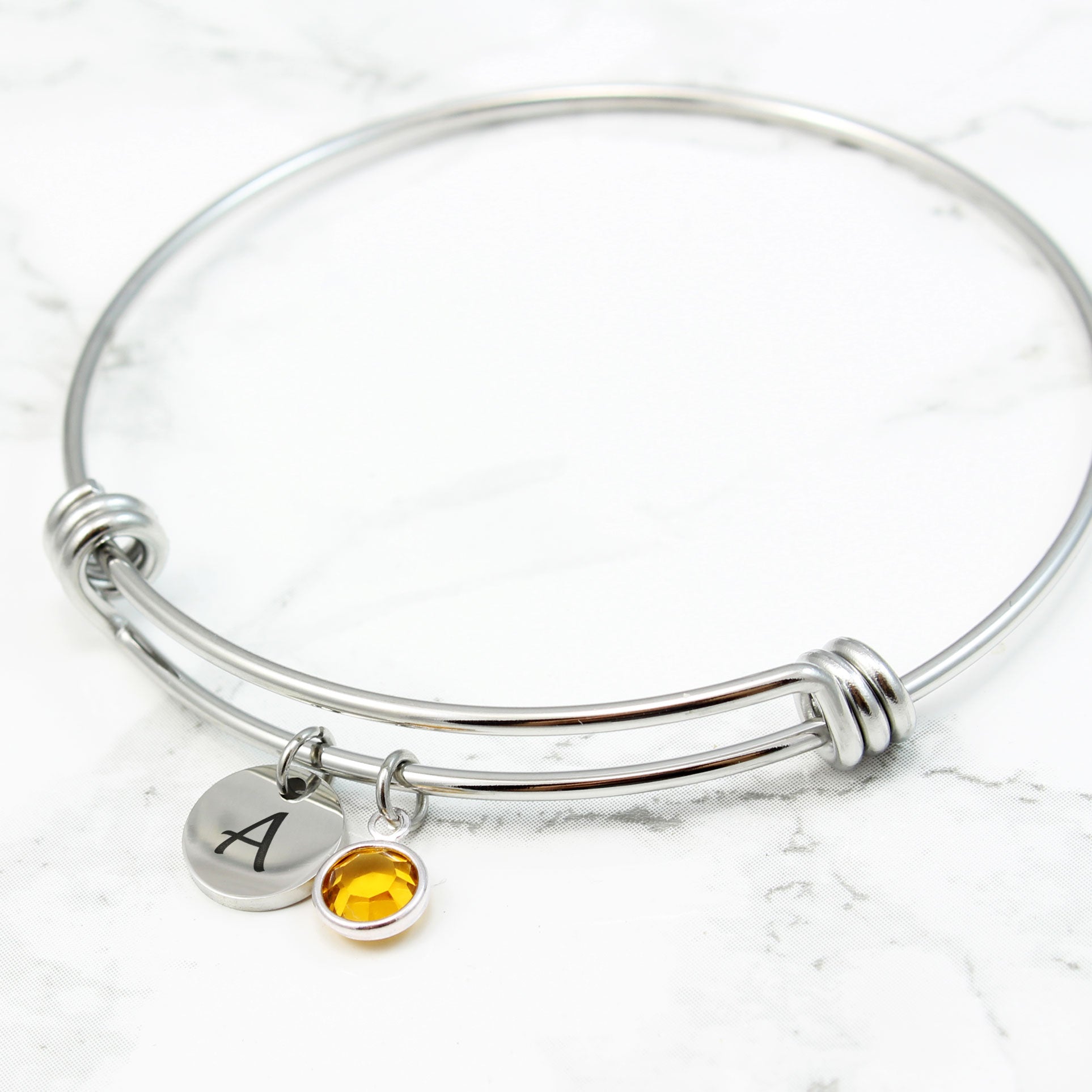 Silver initial and birthstone charm bangle. Adjustable wire bracelet featuring a cursive engraved letter "B" and a yellow-orange gemstone pendant for a personalized touch.