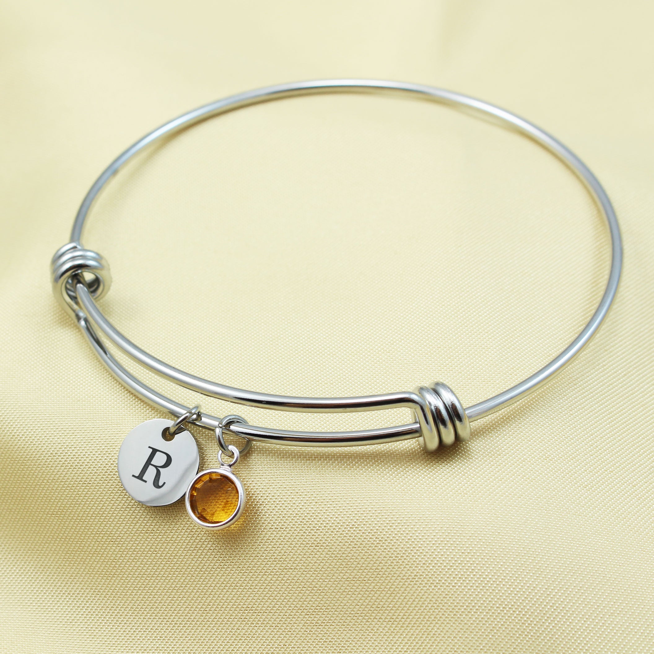 Personalized silver expandable bangle bracelet with a custom engraved initial "B" disc and a round amber birthstone charm. A dainty personalized jewelry gift for birthdays, bridesmaids, or mothers.