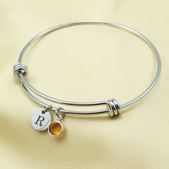 Personalized silver expandable bangle bracelet with a custom engraved initial "B" disc and a round amber birthstone charm. A dainty personalized jewelry gift for birthdays, bridesmaids, or mothers.