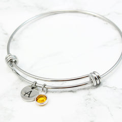 Silver initial and birthstone charm bangle. Adjustable wire bracelet featuring a cursive engraved letter "B" and a yellow-orange gemstone pendant for a personalized touch.