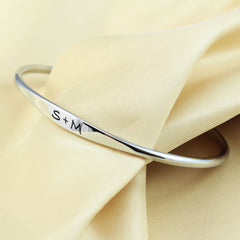 A polished silver bangle bracelet with the engraved initials 'S+M', displayed on draped cream-colored satin fabric.