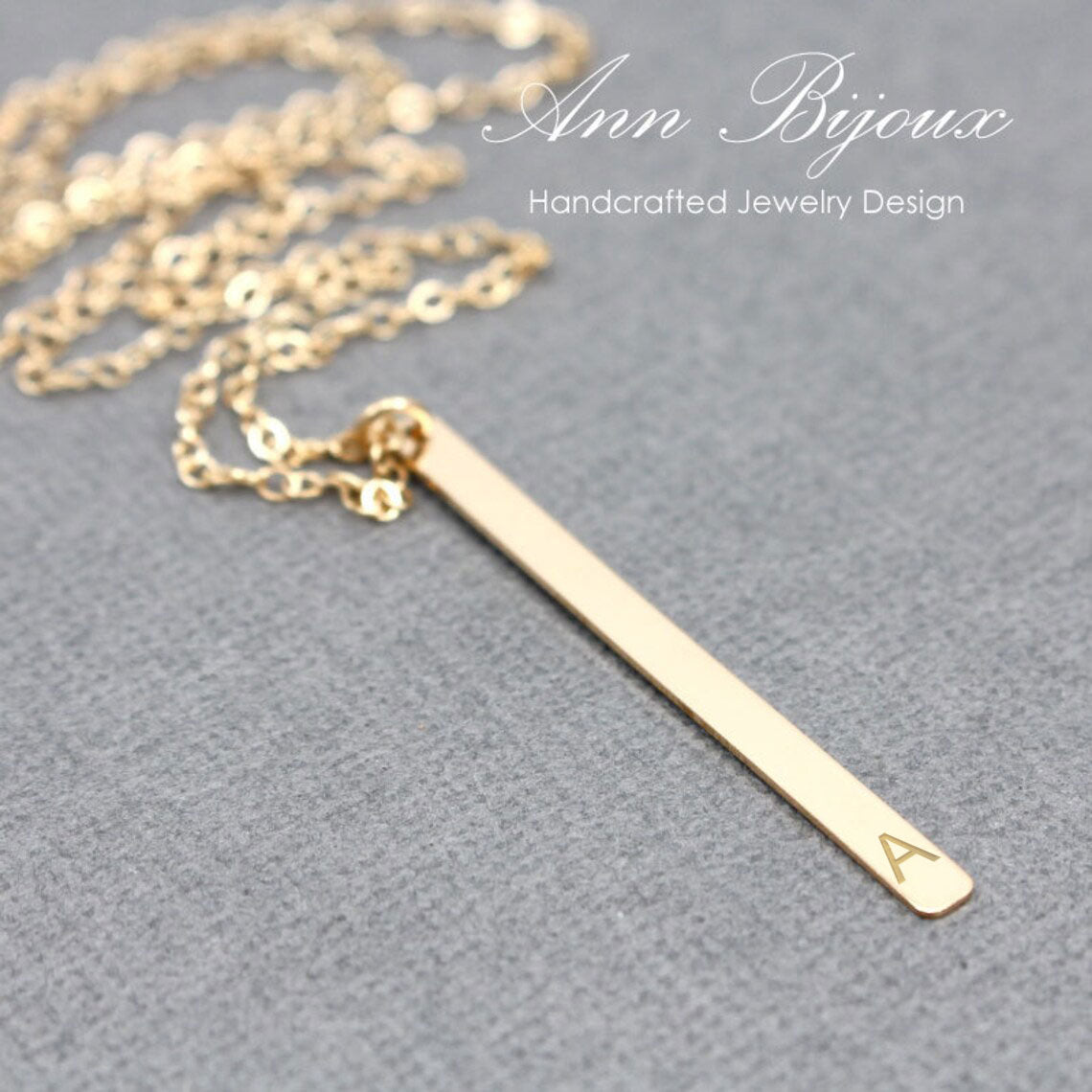 A gold bar necklace with a single letter 'A' engraved at the end, resting on a textured gray surface. The brand name 'Ann Bijoux' is visible in the top right corner.