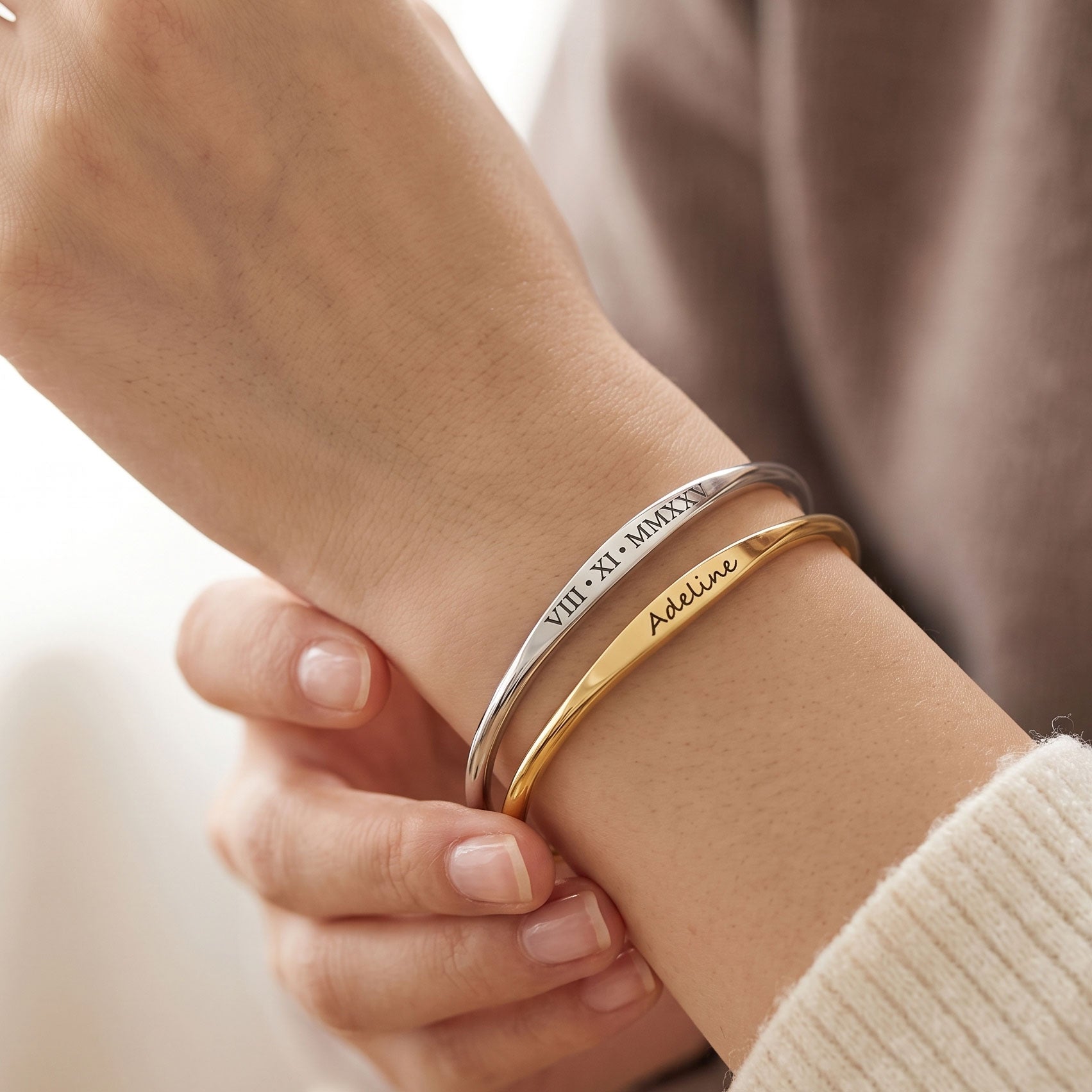 Two slim engraved bangle bracelets on a person's wrist, one silver-toned with Roman numerals and one gold-toned with the name 'Adeline'.