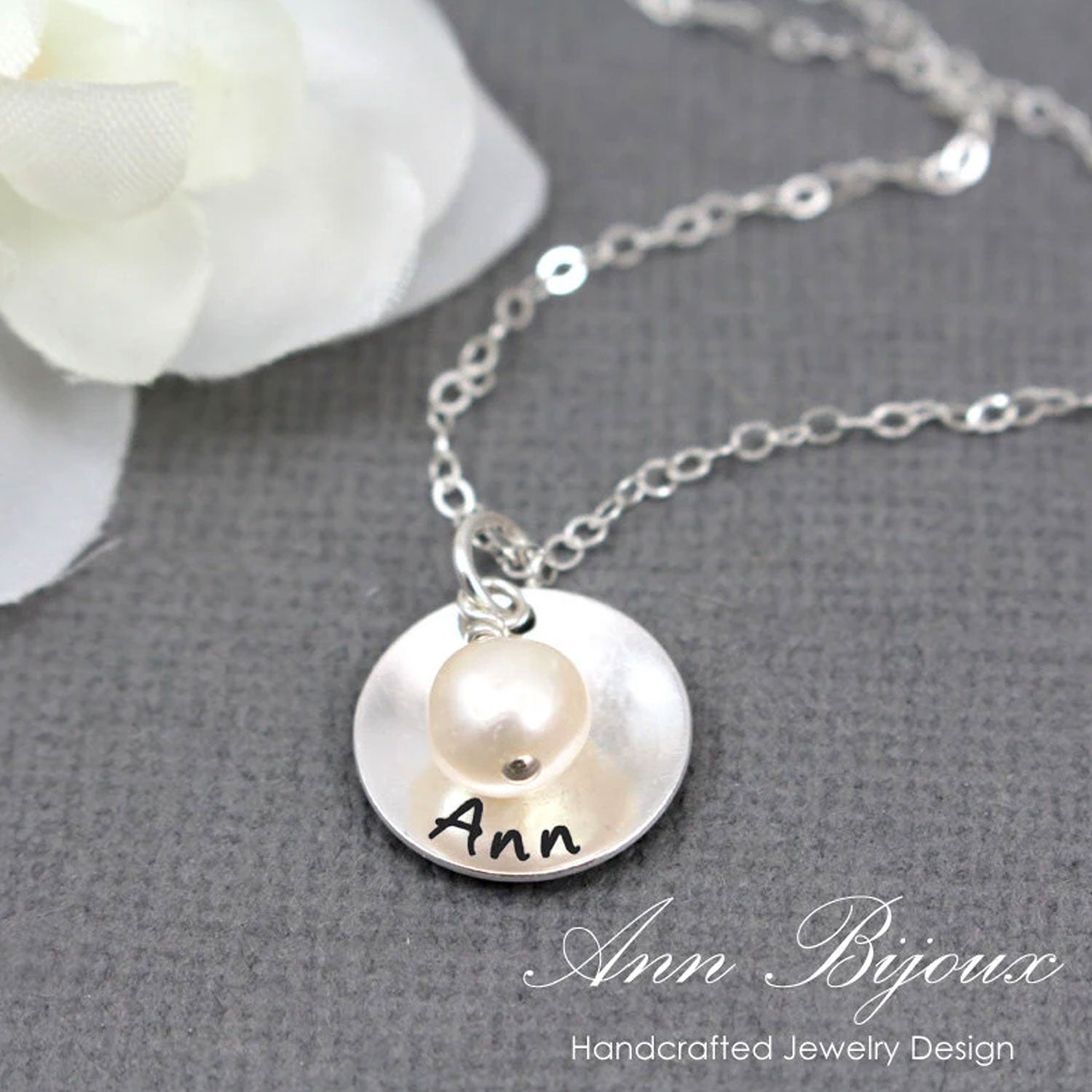 Silver necklace with a round pendant featuring a pearl and engraved name, on a gray surface with a white flower.