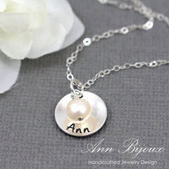 Silver necklace with a round pendant featuring a pearl and engraved name, on a gray surface with a white flower.