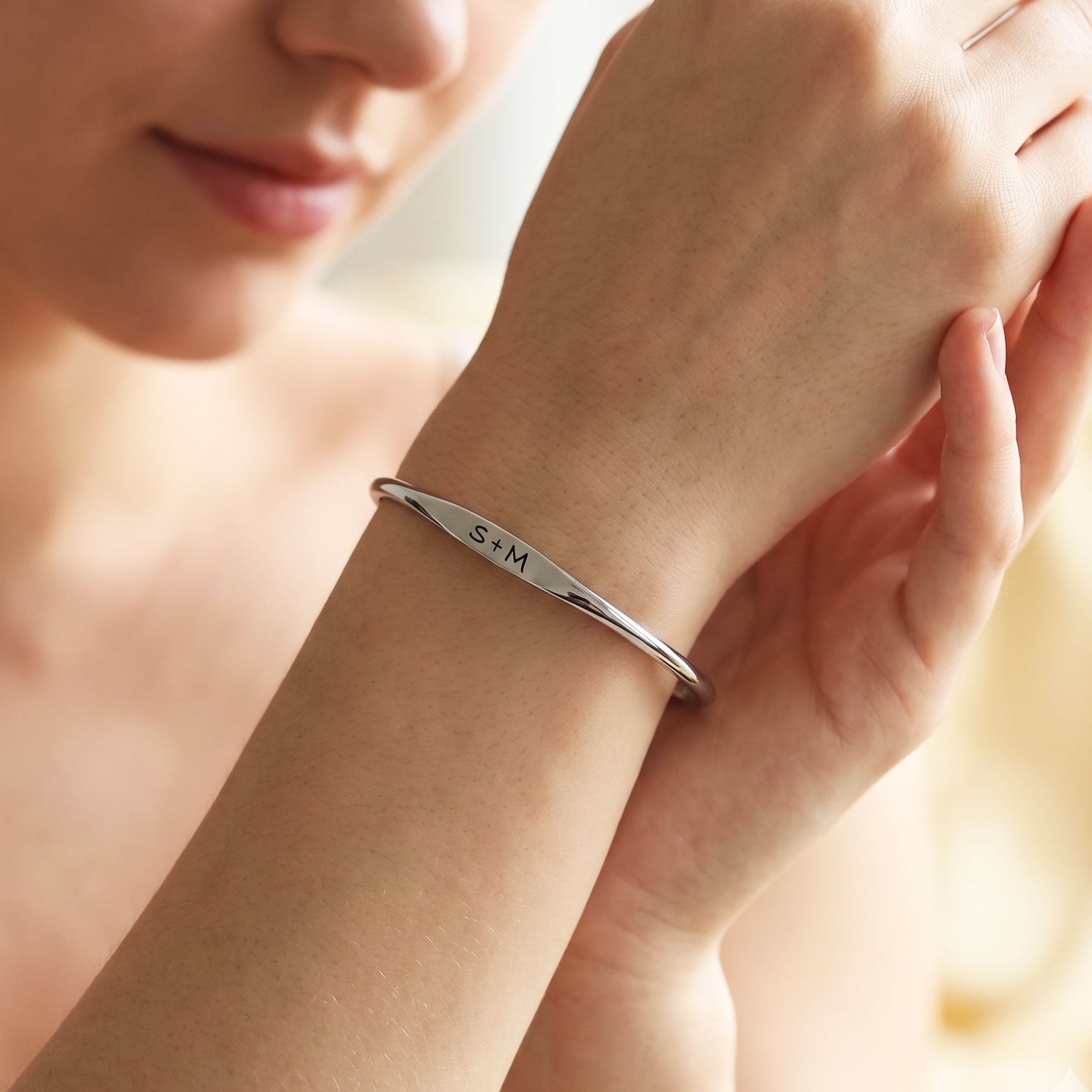 A close-up lifestyle shot of a woman wearing a polished silver bangle bracelet engraved with the initials 'S+M' on her wrist.