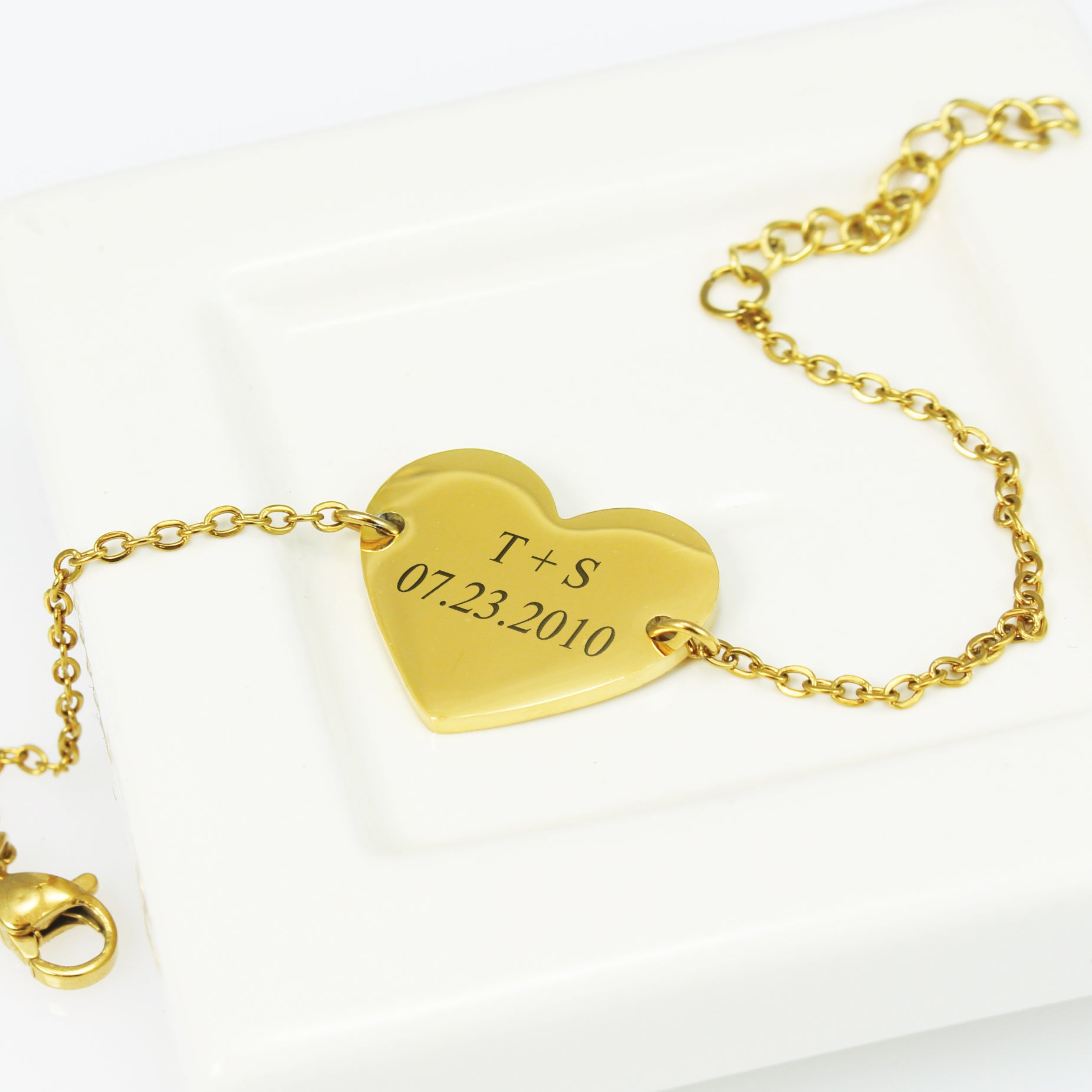 Gold bracelet with a heart-shaped tag engraved with initials and date on a white background