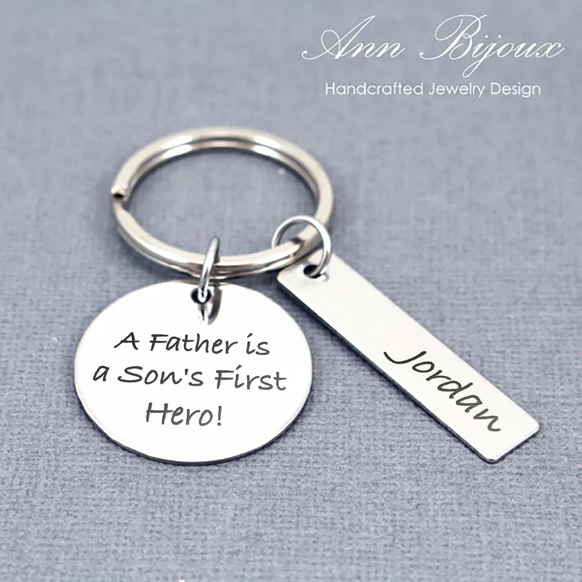 A stainless steel round and bar pendant keychain with an engraved message 'A Father is a Son's First Hero!' along with a name 'Jordan'.