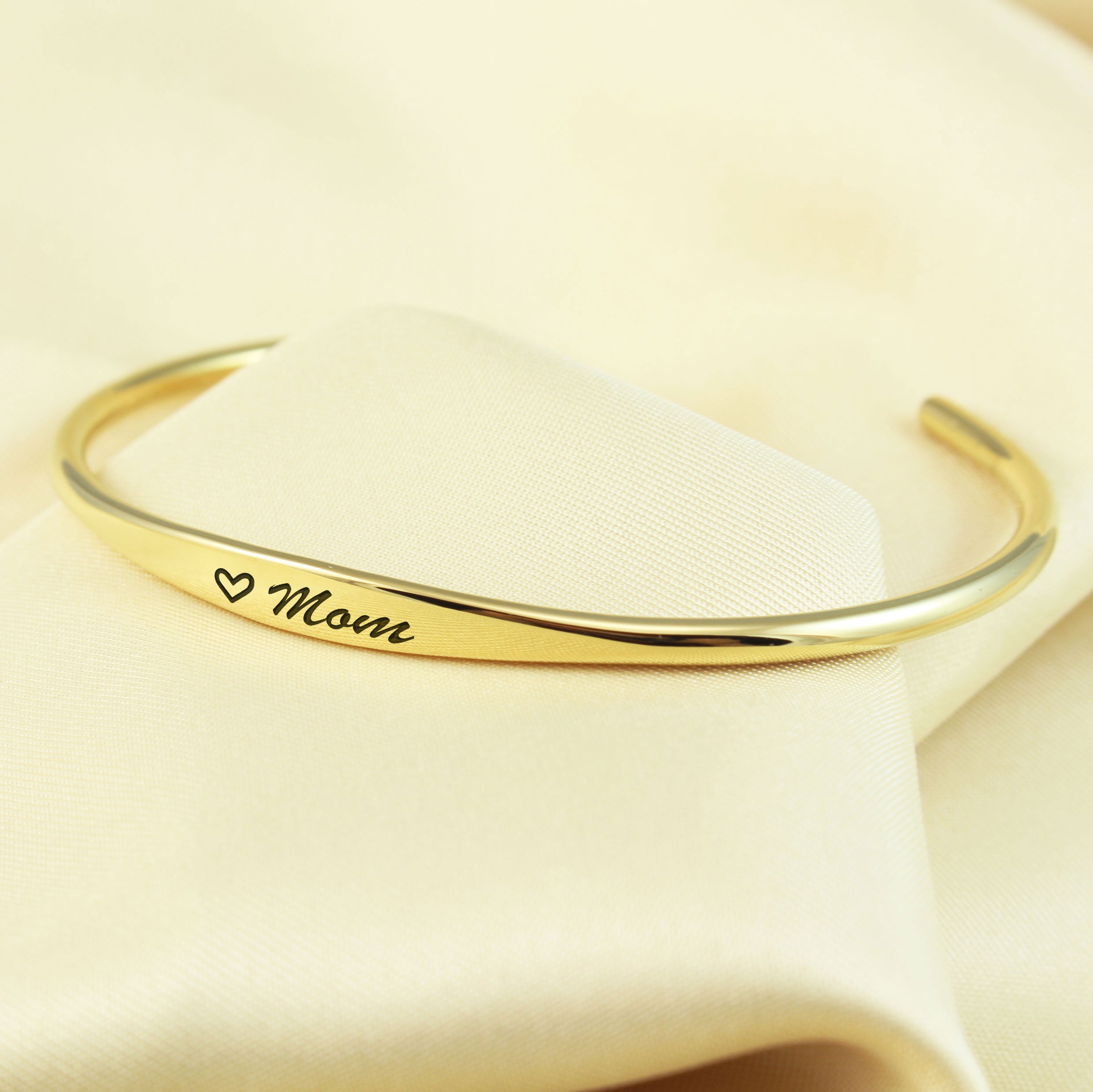 A polished gold cuff bracelet engraved with a small heart and the word 'Mom' in black script, displayed on a soft cream-colored fabric background.