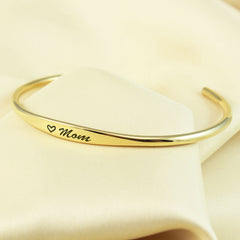 A polished gold cuff bracelet engraved with a small heart and the word 'Mom' in black script, displayed on a soft cream-colored fabric background.