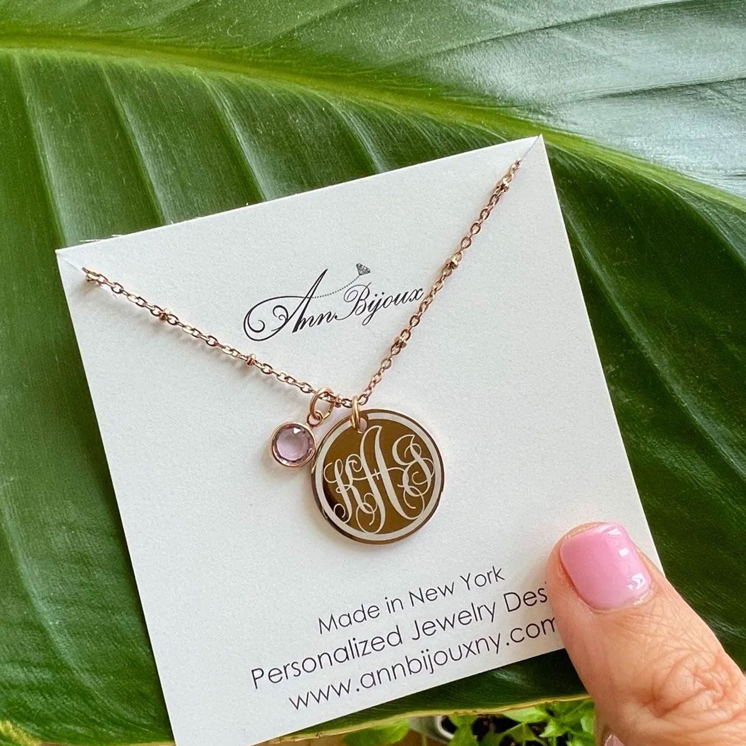 Necklace with monogram pendant on a card, held by a hand against a leafy background