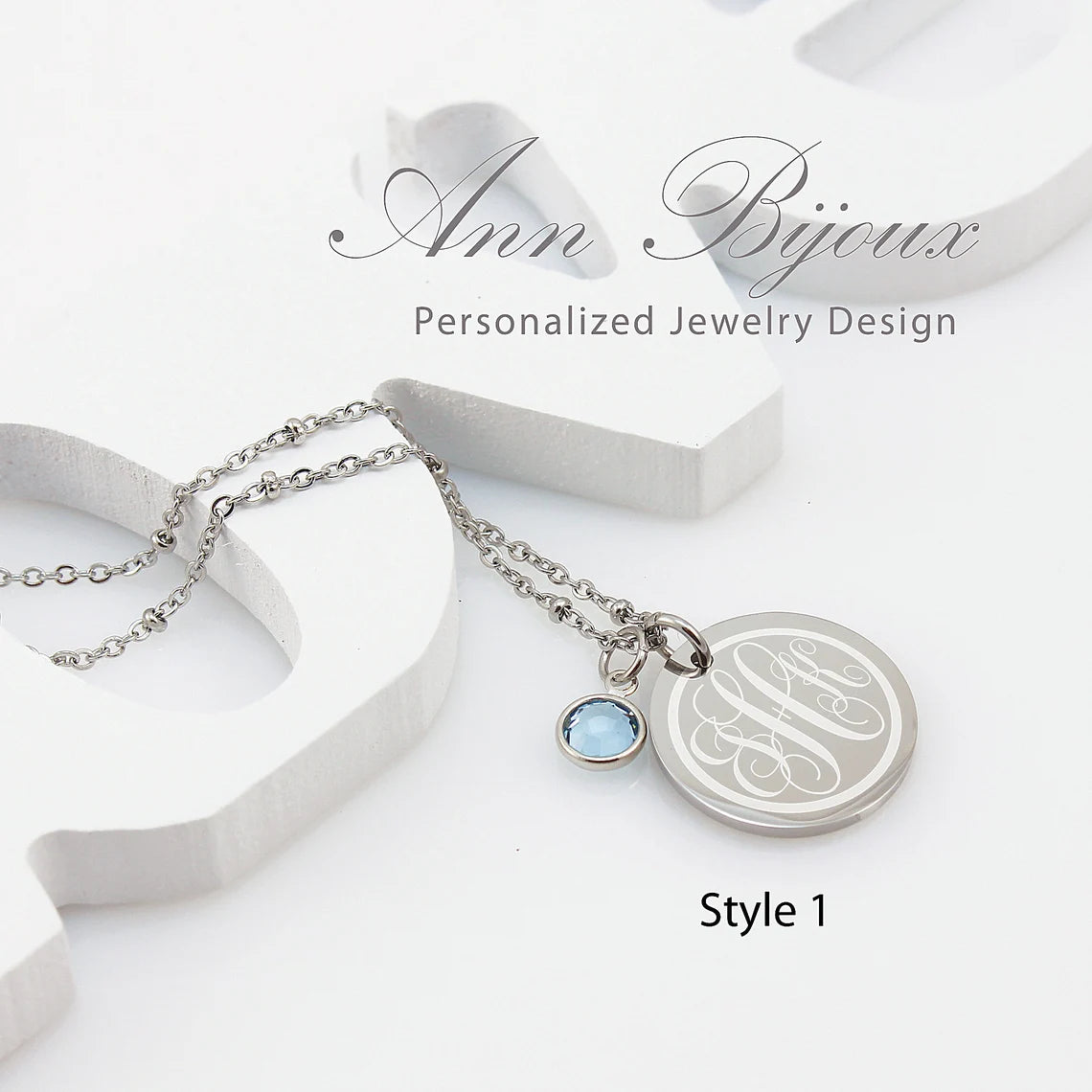 A stainless steel engraved monogram pendant necklace with a birthstone accent, displayed on a white background with the brand name 'Ann Bijoux'.