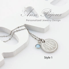 A stainless steel engraved monogram pendant necklace with a birthstone accent, displayed on a white background with the brand name 'Ann Bijoux'.