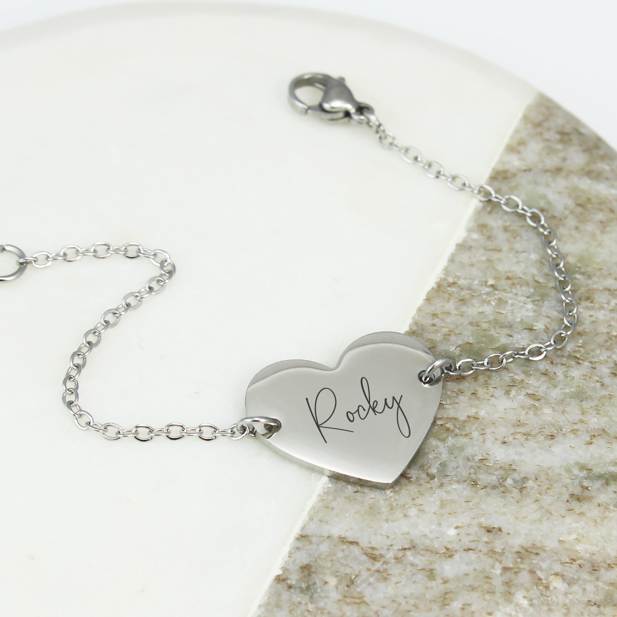 Silver bracelet with a heart-shaped tag engraved with 'Rocky' on a stone surface