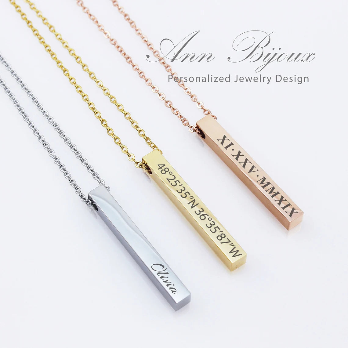 Three vertical 3D bar necklaces in silver, gold, and rose gold finishes. The silver bar is engraved with the name 'Olivia,' the gold bar features geographic coordinates, and the rose gold bar shows a date in Roman numerals.