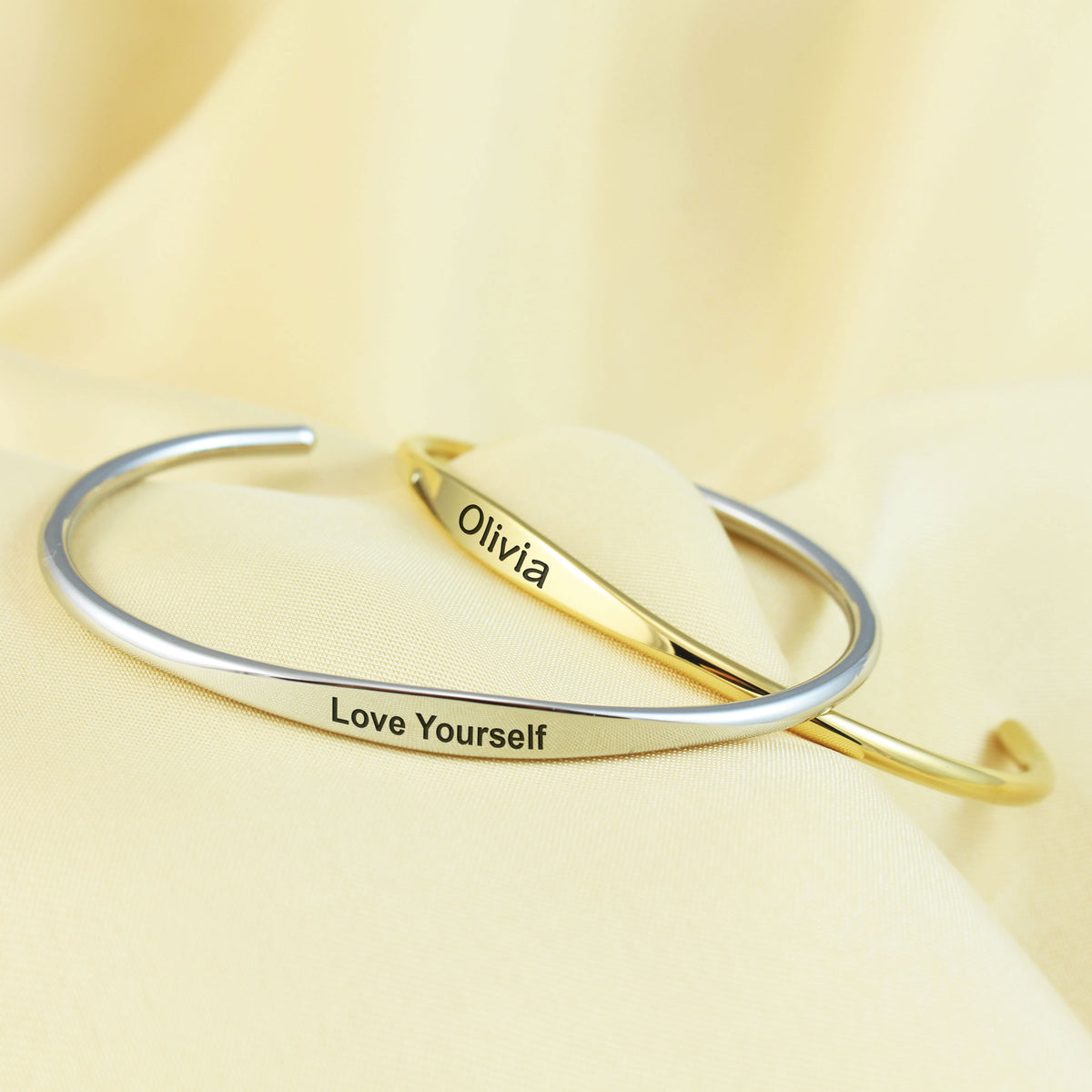 Two slim open cuff bracelets, one silver-toned and one gold-toned, displayed on a soft cream fabric background. The silver bracelet is engraved with 'Love Yourself', and the gold bracelet is engraved with 'Olivia'.