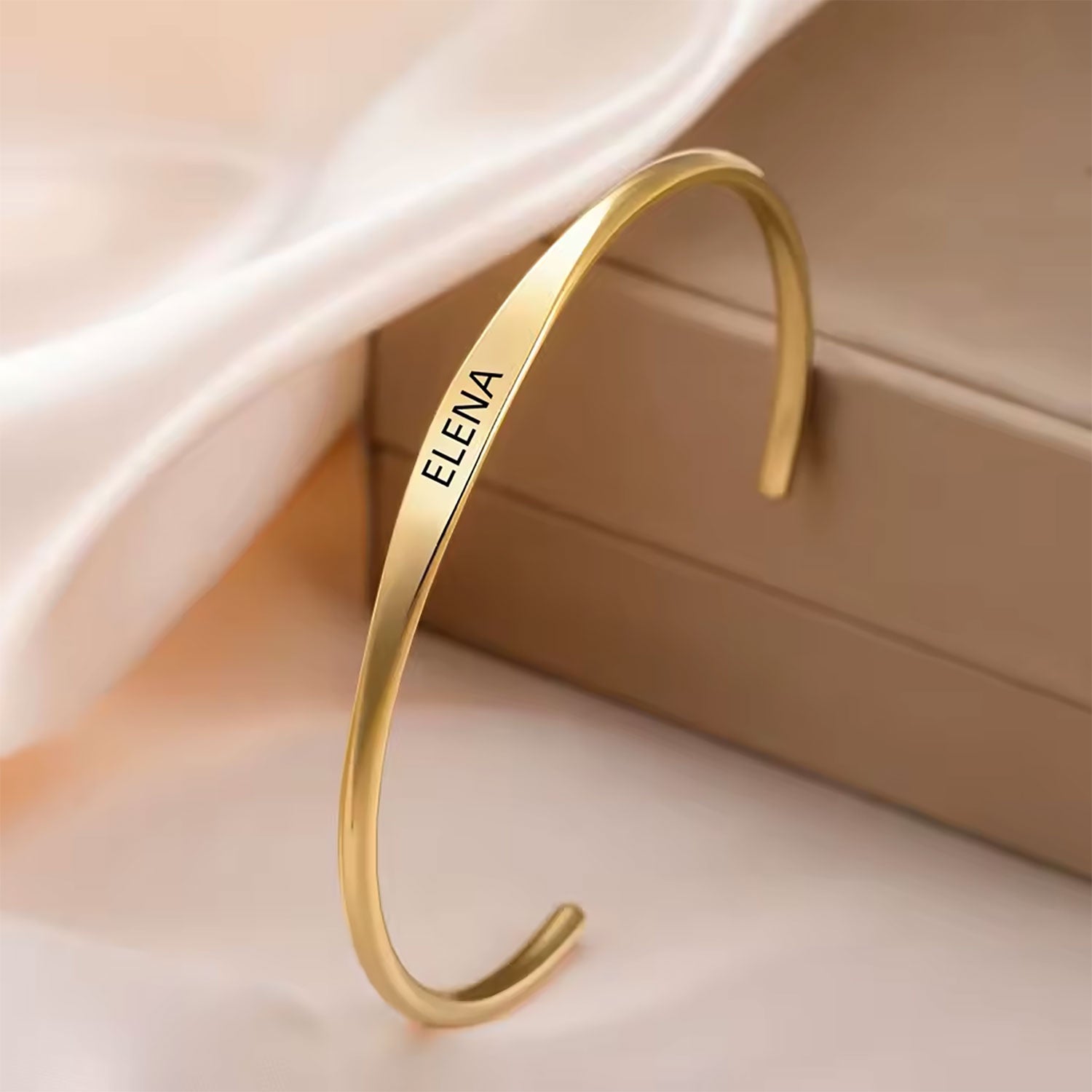 A slim gold-tone open cuff bangle bracelet with the name 'ELENA' engraved on the front, displayed on a beige jewelry box with soft white fabric in the background.