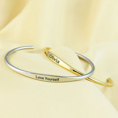 Two slim open cuff bracelets, one silver-toned and one gold-toned, displayed on a soft cream fabric background. The silver bracelet is engraved with 'Love Yourself', and the gold bracelet is engraved with 'Olivia'.