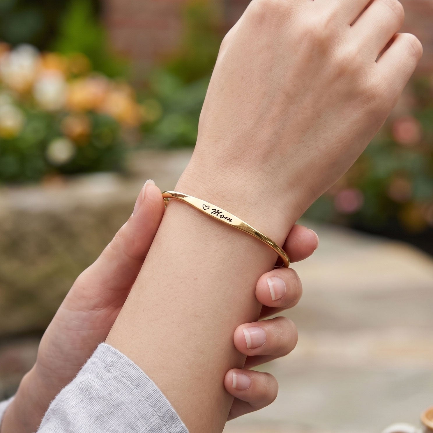 A gold bangle bracelet engraved with 'Mom' and a heart, worn on a person's wrist and held by another hand, with a blurred outdoor garden background.