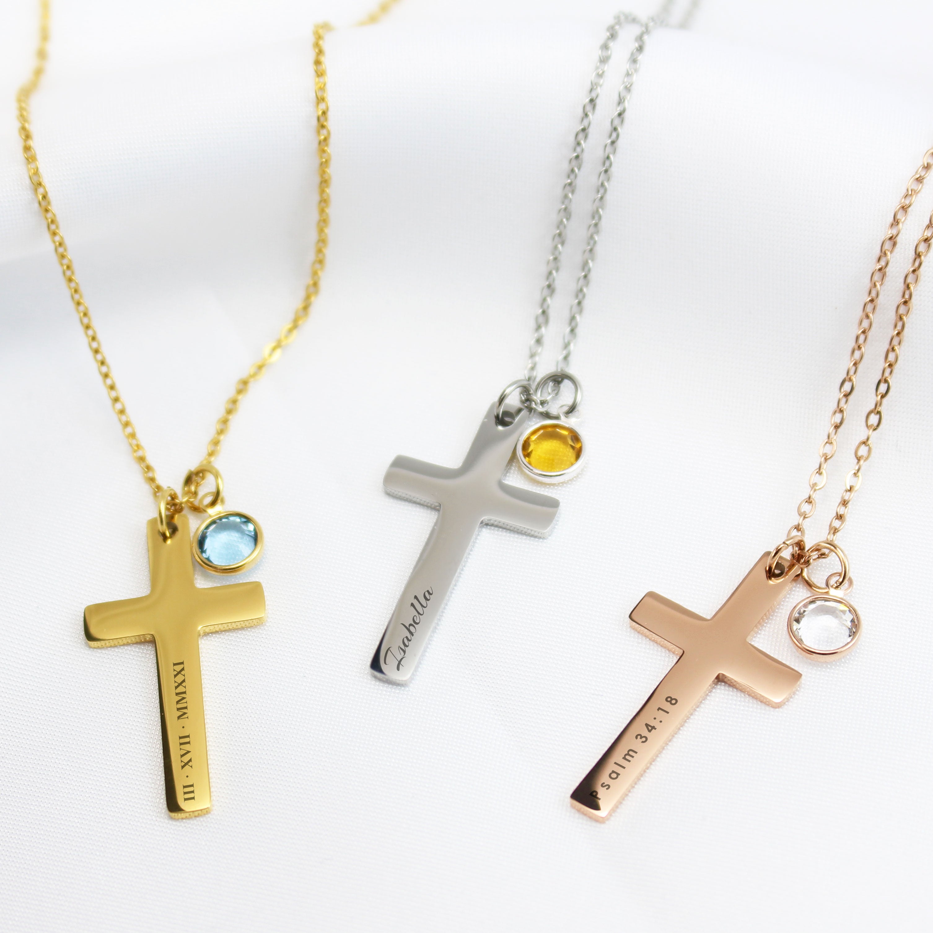 Three personalized cross necklaces in gold, silver, and rose gold. Each cross features a different custom engraving—Roman numerals, the name Isabella, and a Bible verse—paired with a small, colorful round birthstone charm.