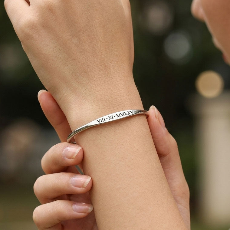 A close-up of a person's wrist wearing a slim silver bangle bracelet with engraved Roman numerals, with their other hand holding the wristband in place against a softly blurred outdoor background.