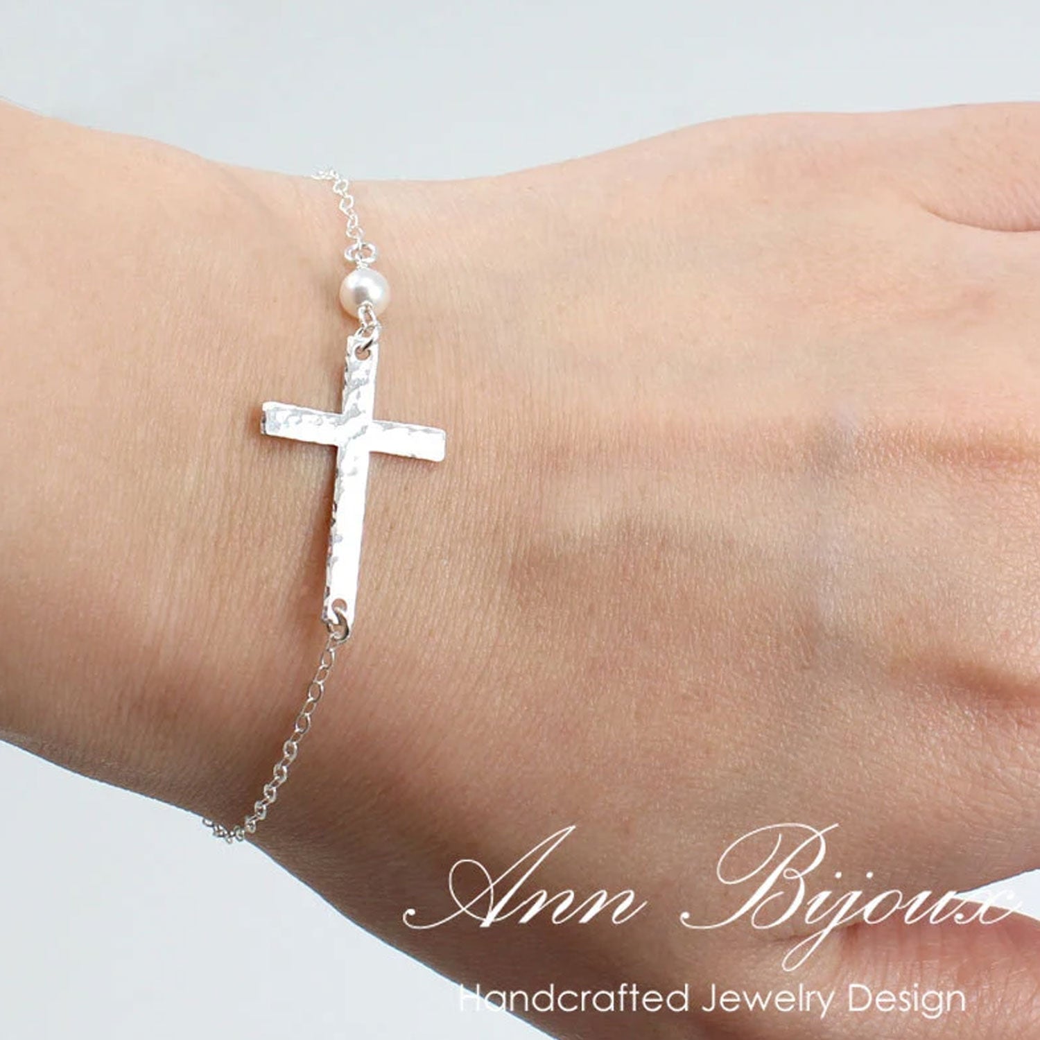Silver cross bracelet with a pearl on a wrist, branded 'Ann Bijoux'.