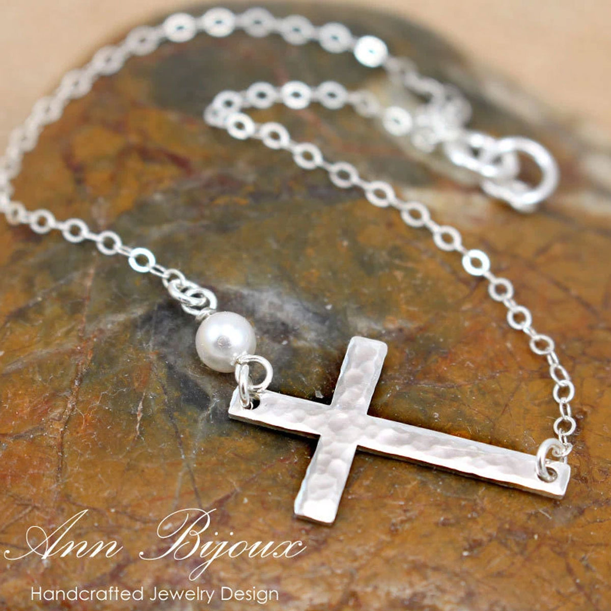 Silver bracelet with a cross pendant and pearl on a stone surface, branded 'Ann Bijoux'.