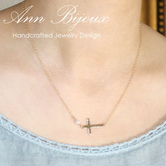 Close-up of a horizontal gold cross with a textured, hammered surface connected to a single white pearl on a gold chain, resting on a neutral fabric background.