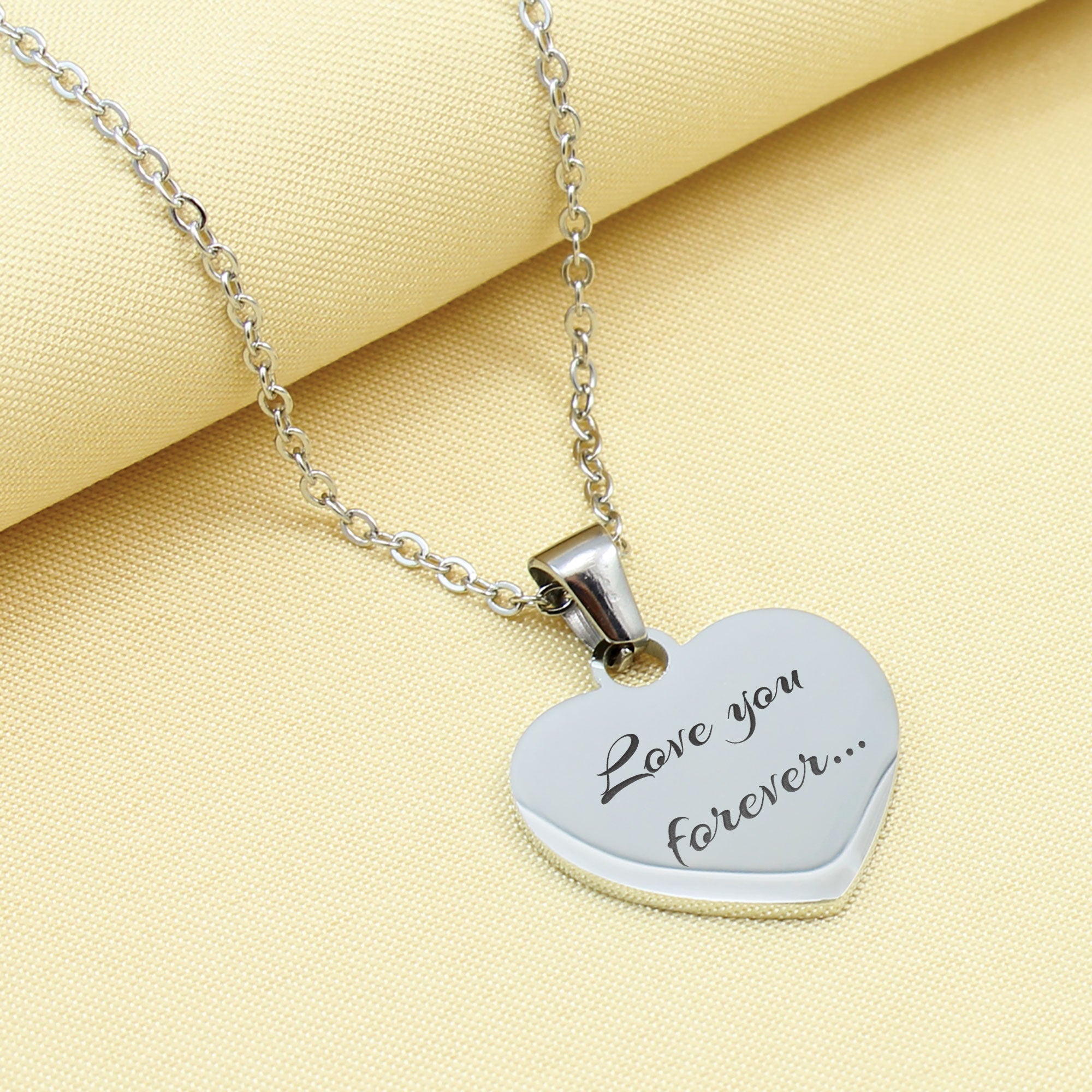 Heart-shaped silver pendant with 'Love you forever...' engraving on a yellow fabric background